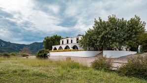 Un edificio bianco con finestre ad arco e un balcone, circondato da vegetazione e montagne.