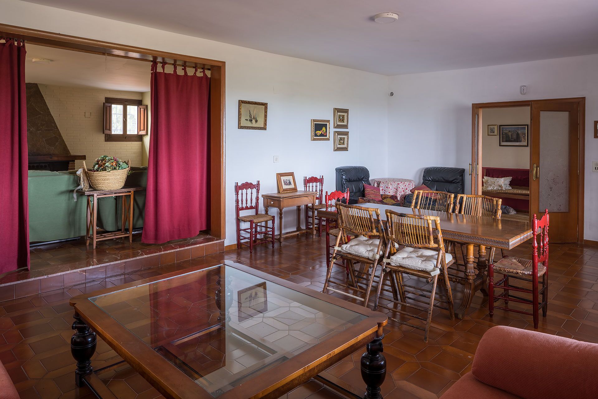 Un coin repas avec une table à plateau en verre et des chaises en bois, un coin cuisine avec un rideau rouge, et un salon avec un canapé et un fauteuil rouge.