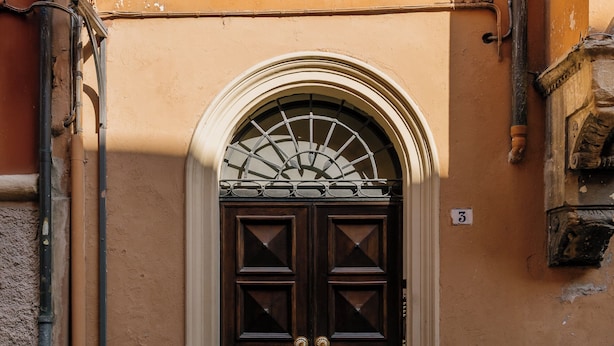 Una puerta marrón con un ventanal decorativo en la parte superior, flanqueada por dos pilares y una farola a la izquierda.
