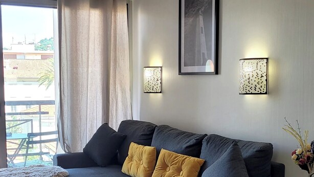 Een moderne woonkamer met een donkergrijze modulaire bank, twee gele kussens, een klein houten salontafel en een bijzettafeltje. Er zijn twee wandlampen en een ingelijst schilderij boven de bank.