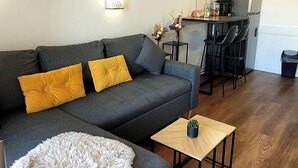 Een moderne woonkamer met een grijze bank, gele kussens, een pluizig witte poef en een houten salontafel. Op de achtergrond is een keukenhoek met barstoelen en een hanglamp zichtbaar.