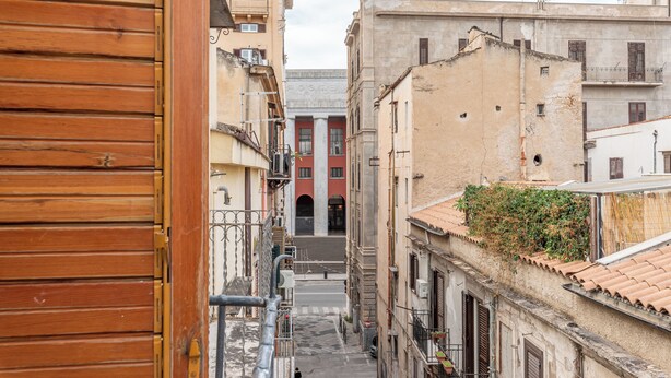 Uma rua estreita com edifícios de ambos os lados, um marco de porta de madeira à esquerda e uma escadaria que leva à entrada de um edifício.