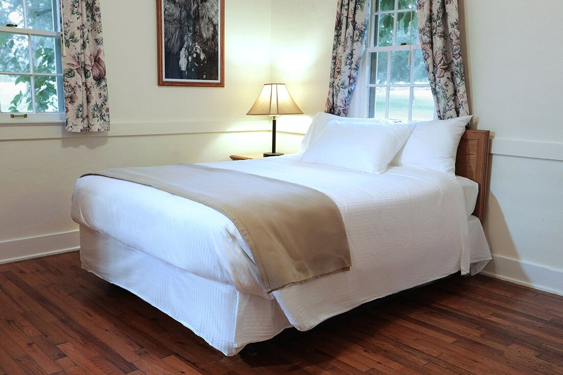Un lit bien fait, avec du linge de lit blanc et un plaid beige, une lampe de chevet et des rideaux à motifs floraux à une fenêtre.