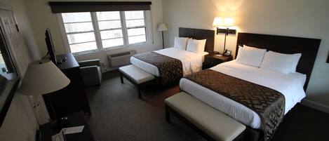 A hotel room with two beds, a desk, a chair, a lamp, and a window.