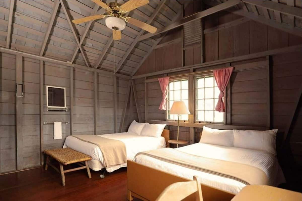 Une pièce rustique en bois, avec deux lits, un ventilateur de plafond et une fenêtre avec des rideaux.