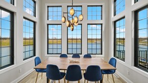 A dining room with a large wooden table, six blue chairs, and a chandelier. The room has large windows with a view of a body of water and a grassy field. - La Dolce Vita (Ottawa)
