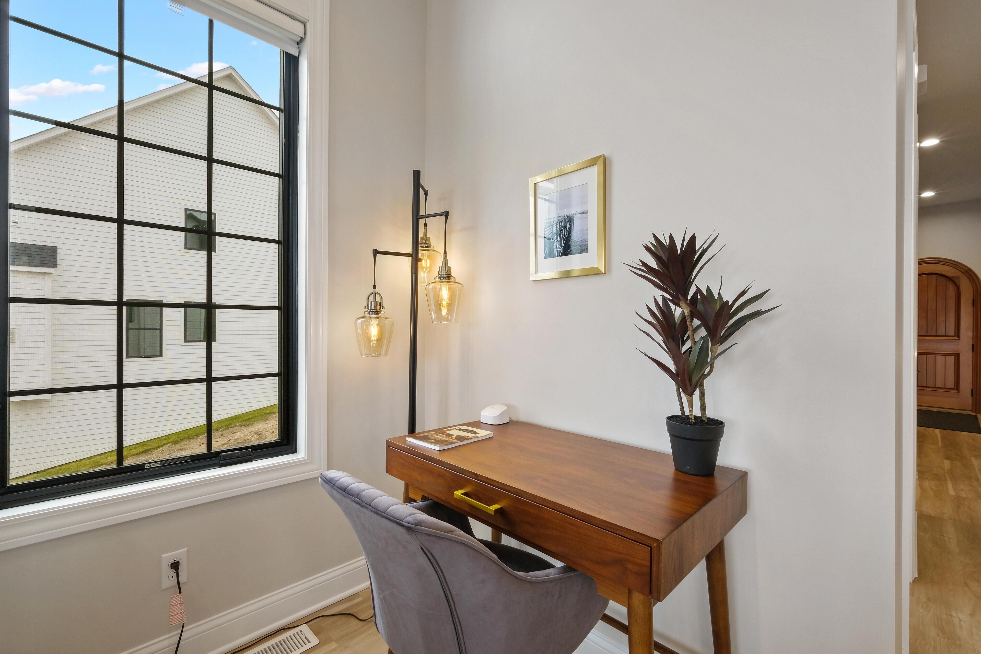 A room with a large window, a desk with a chair, a potted plant, a framed picture, and a wall-mounted light fixture.