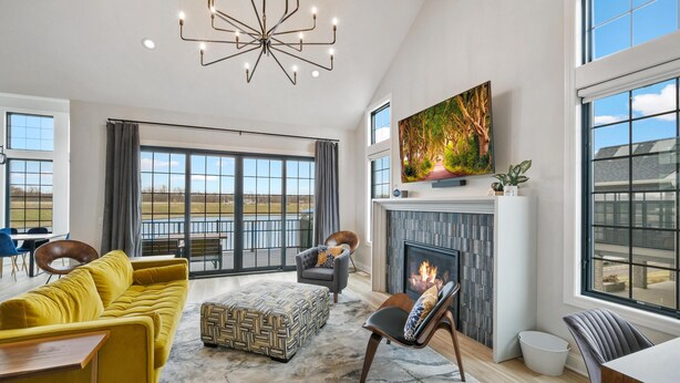 A spacious living room with a yellow sofa, a patterned ottoman, a fireplace, and a large window with a view of the outdoors. - La Dolce Vita (Ottawa)
