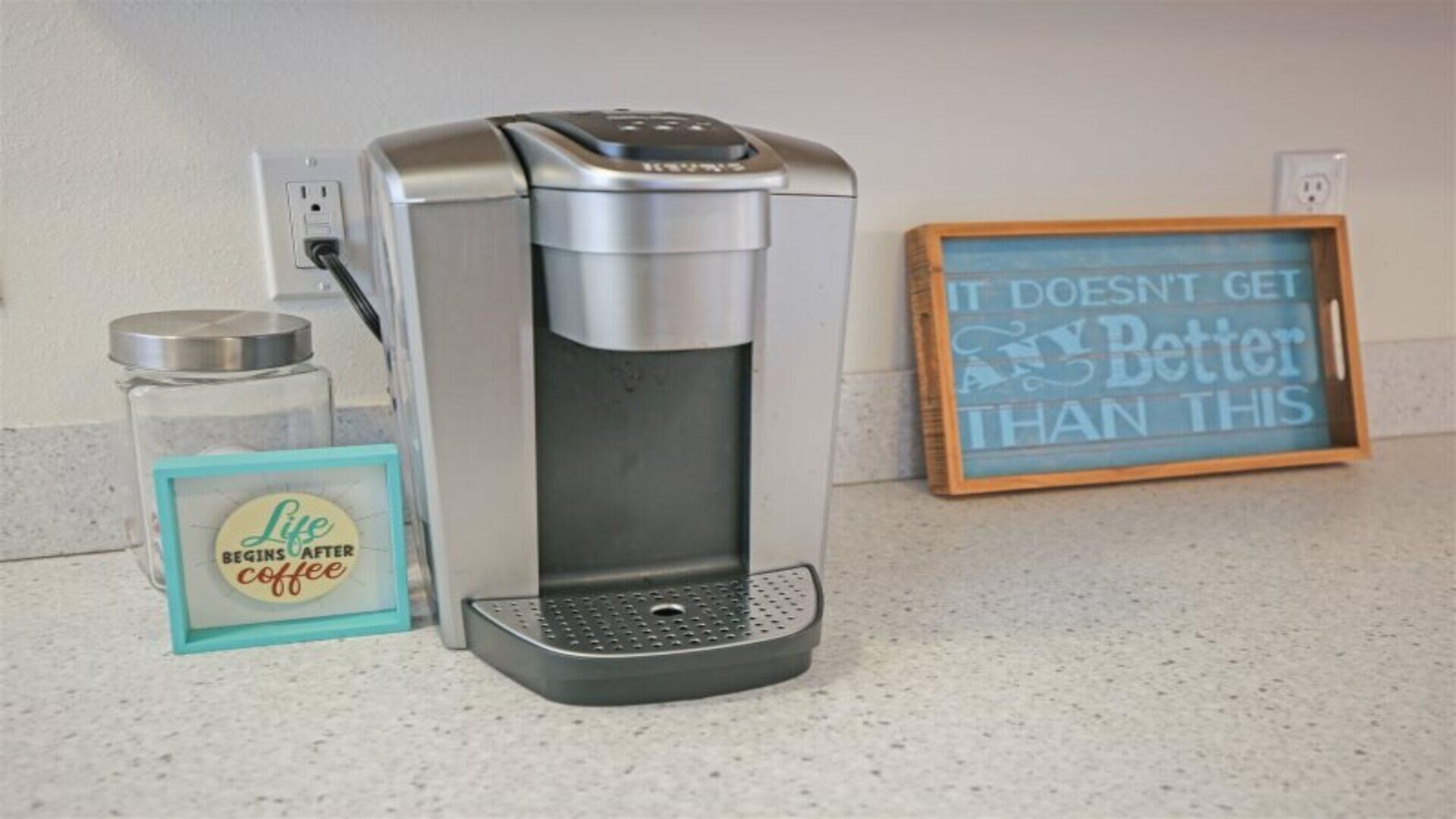 A coffee maker on a countertop next to a jar labeled "Life Begins After Coffee" and a framed sign that reads "It doesn't get better than this."