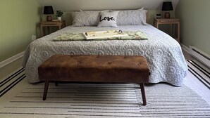 Un dormitorio con una cama, un banco, dos mesitas de noche y un cuadro colgado en la pared.