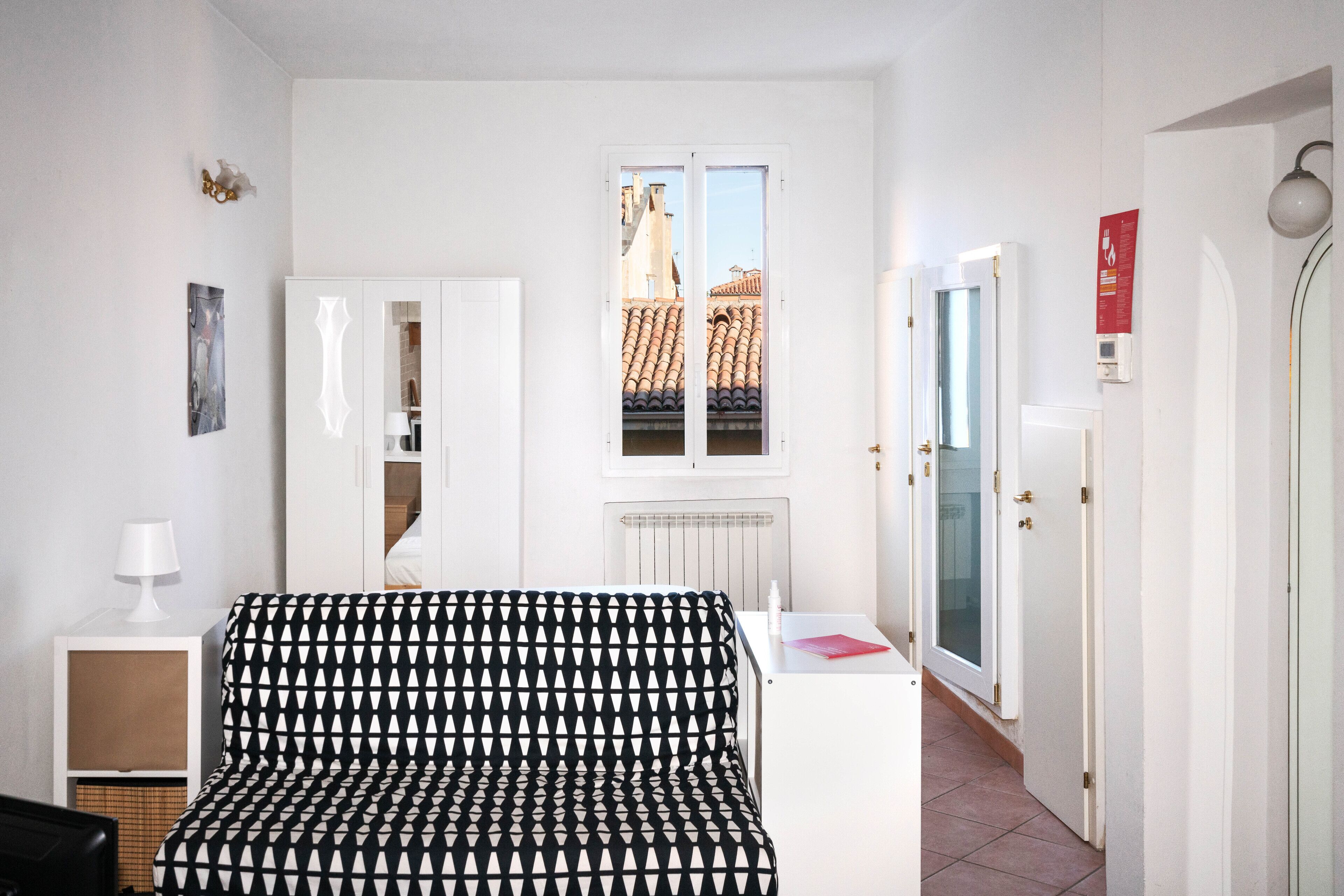 Une chambre d’hôtel moderne avec un canapé à motifs, une petite table avec une lampe, un radiateur et une vue sur le paysage urbain par la fenêtre.
