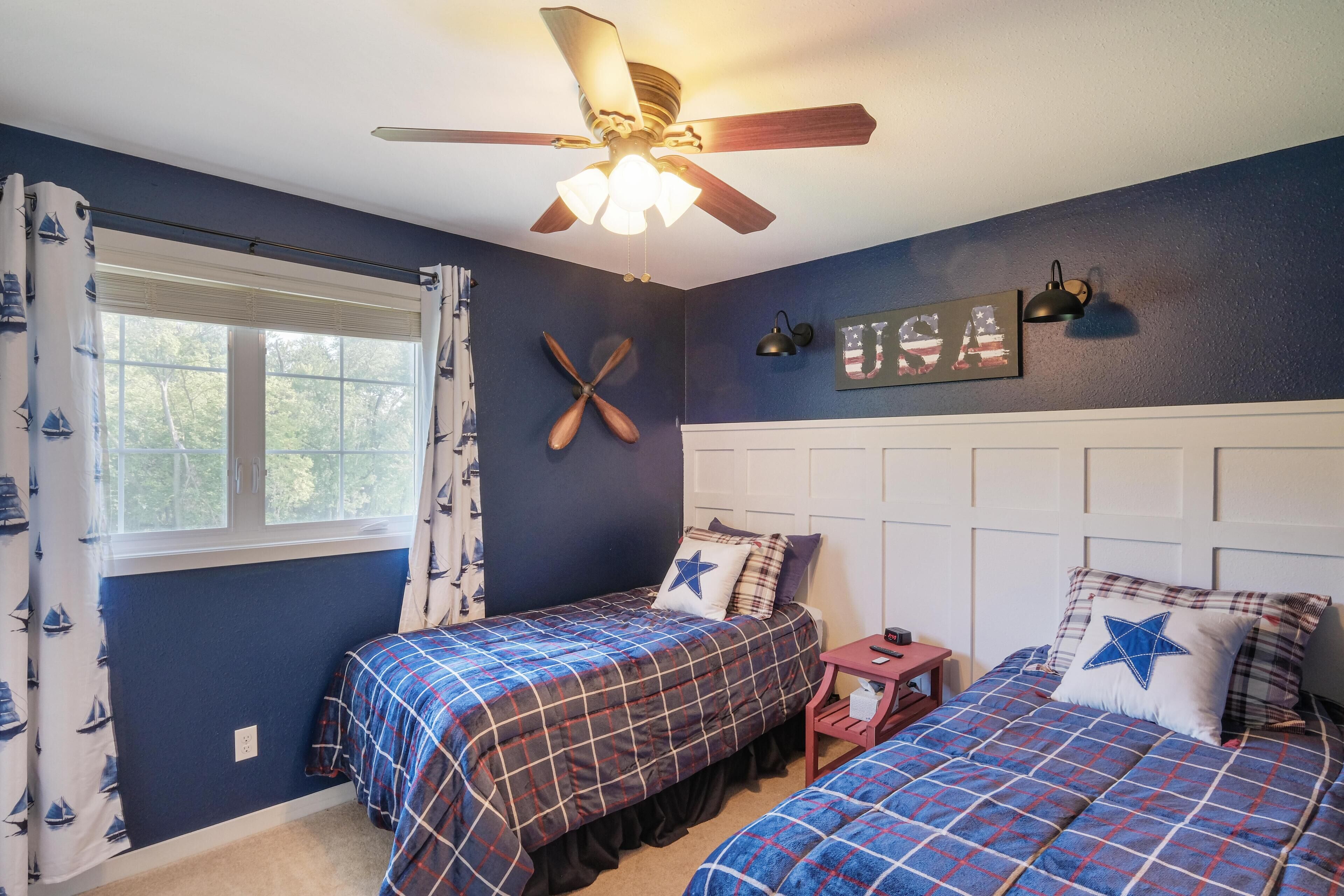 A bedroom with two beds, a ceiling fan, a window with curtains, and a wall-mounted light fixture.