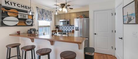 A kitchen with a bar area, a ceiling fan, and a door with a painting.