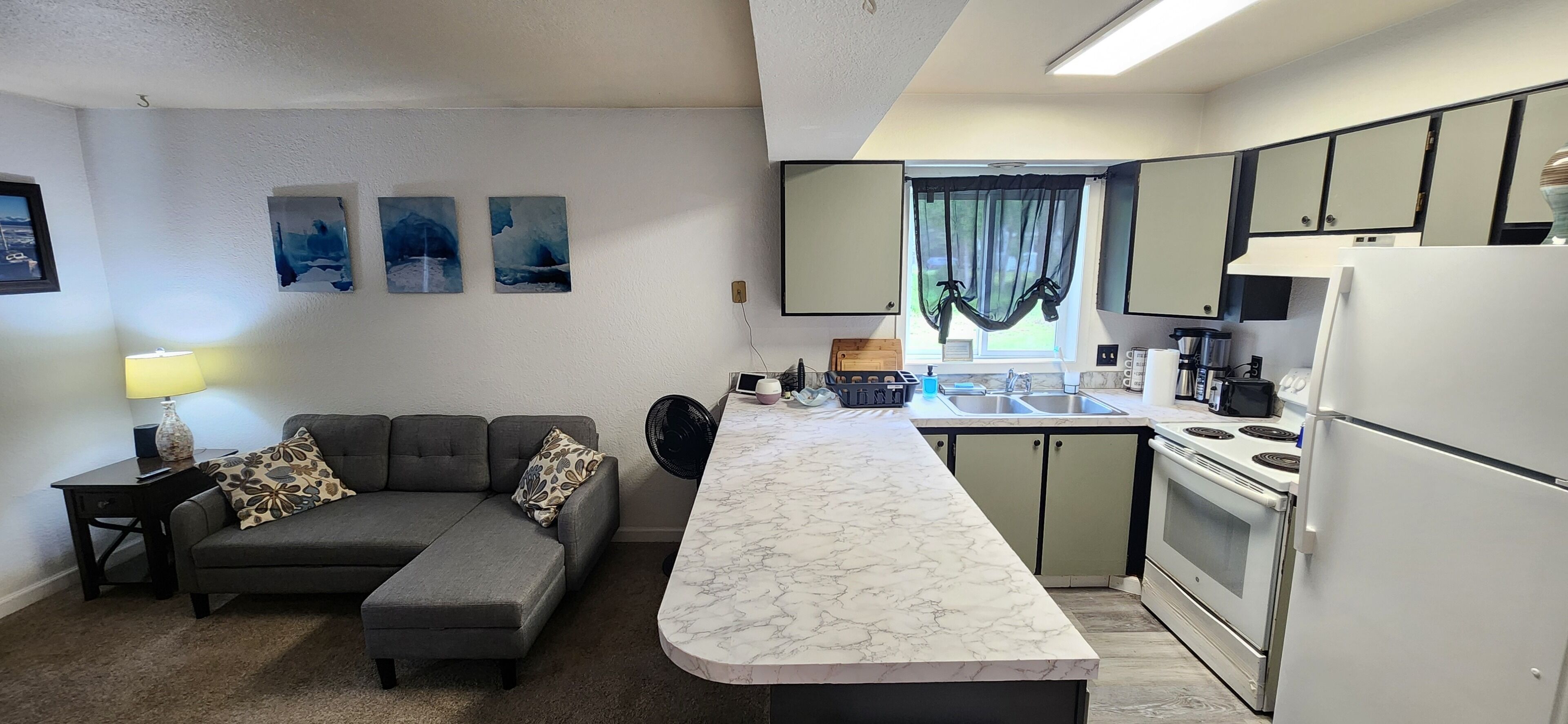 A compact living space with a sofa, a dining table, a kitchen area including a stove and refrigerator, and artwork on the walls.
