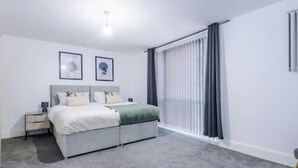 A modern bedroom with a large bed, a bedside table, a radiator, and a window with curtains. - Old Dutch Embassy by Guestz (Kingston-upon-Hull)