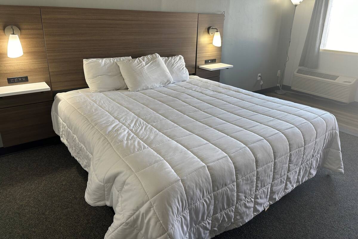 A neatly made bed with a white quilt, wooden headboard, bedside lamps, and a window with curtains.