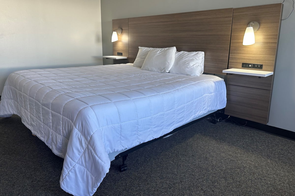 A hotel room with a large bed, two bedside tables with lamps, a wooden headboard, and a carpeted floor.