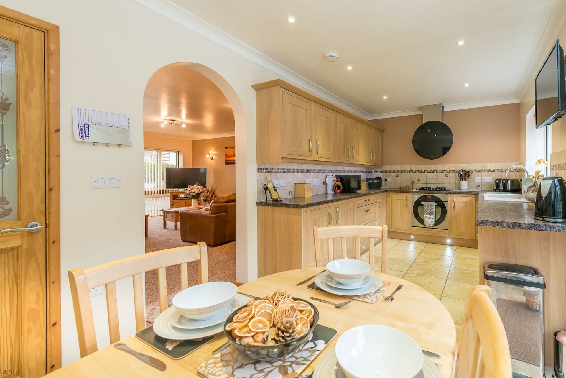 A modern kitchen with wooden cabinets, a dining area with a table set for two, and a TV in the living room.