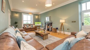 A spacious living room with a fireplace, leather sofas, and a large window. - Beautiful Farmhouse Accommodation in a Country Setting | Hendre-Feinws (PWLLHELI)