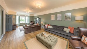 A spacious living room with a dark brown leather sofa, a green ottoman, and a grey sectional sofa. There are framed artworks on the walls and a chandelier above the seating area. - Large Detached Property Walking Distance To The Beach | Llywelyn-Fawr (Aberffraw)