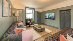 A spacious living room with a grey sofa, a brown leather couch, and a grey ottoman. There is a television mounted on the wall, a chandelier, and a window providing natural light. - Large Detached Property Walking Distance To The Beach | Llywelyn-Fawr (Aberffraw)