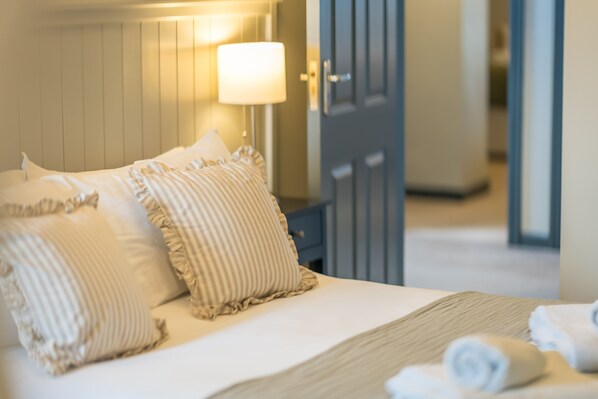 A neatly made bed with striped pillows and a rolled towel, a bedside lamp, and a door in the background. - Large Detached Property Walking Distance To The Beach | Llywelyn-Fawr (Aberffraw)