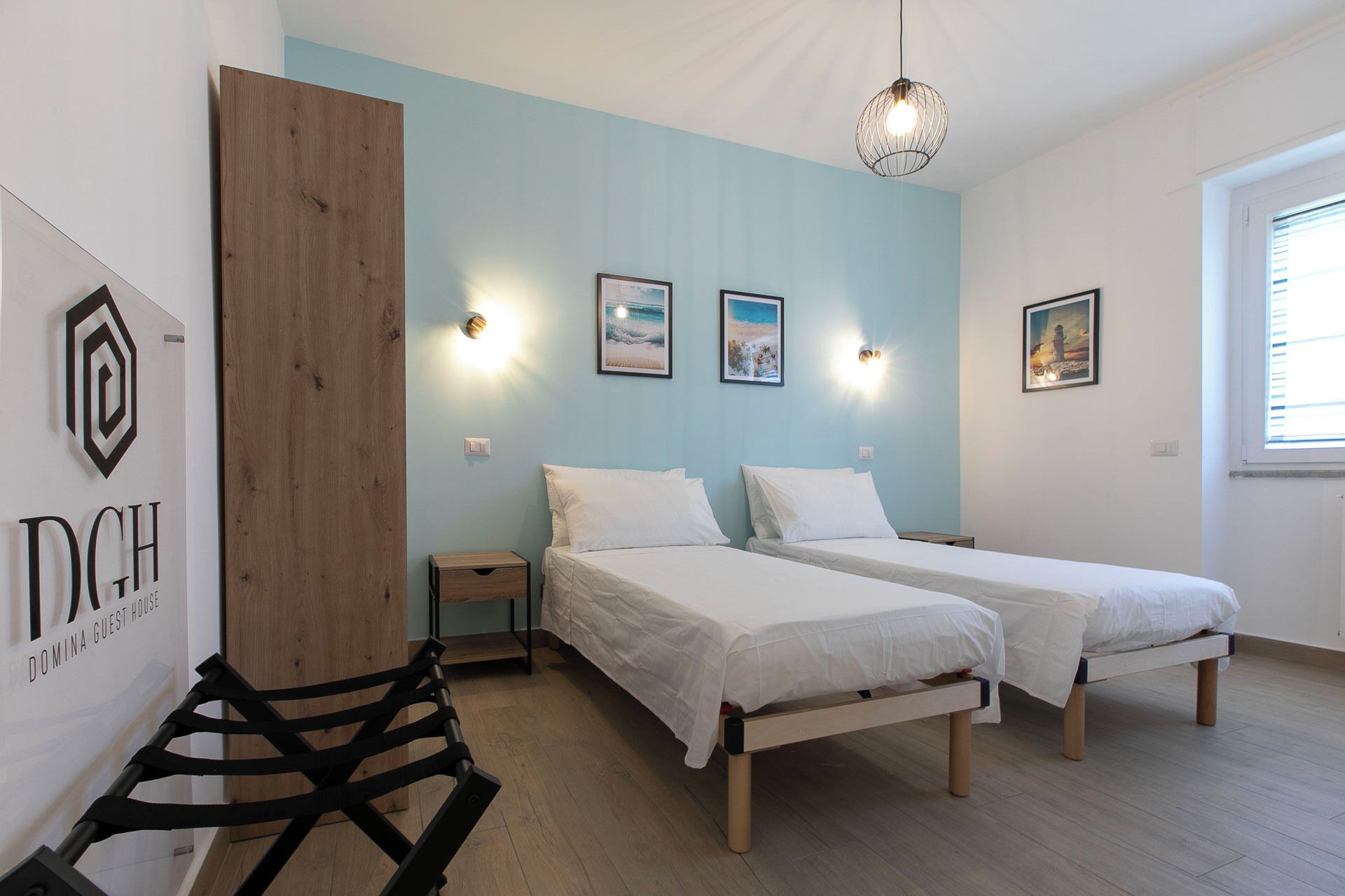 A guest room with two beds, a wooden desk, a chair, and a wall-mounted light fixture.