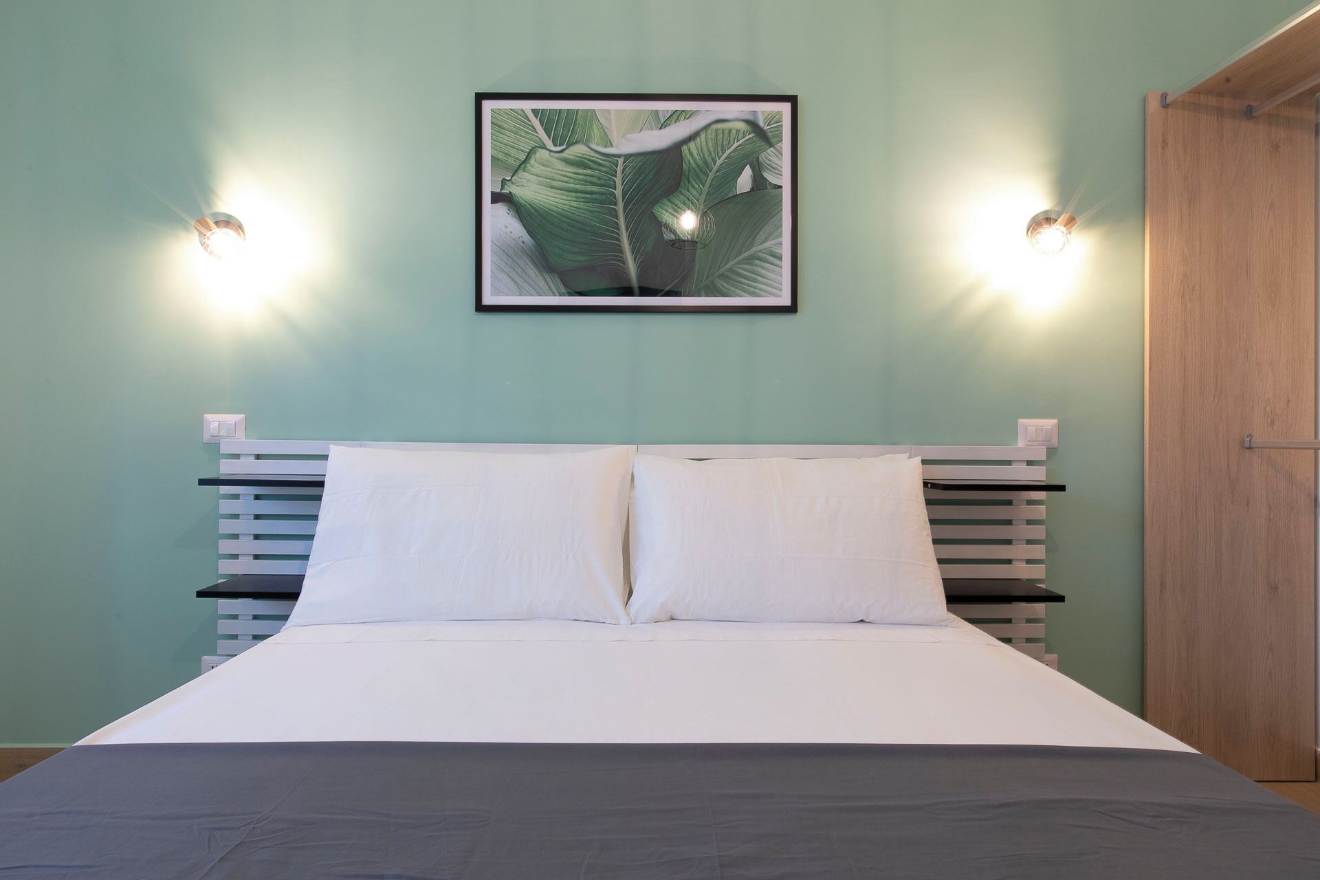 A bed with white bedding, a framed picture above it, and two wall-mounted lights.