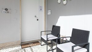 Entrada de hotel con una puerta blanca, dos sillones de mimbre, una mesita con un jarrón rosa y dos macetas colgantes con plantas verdes.