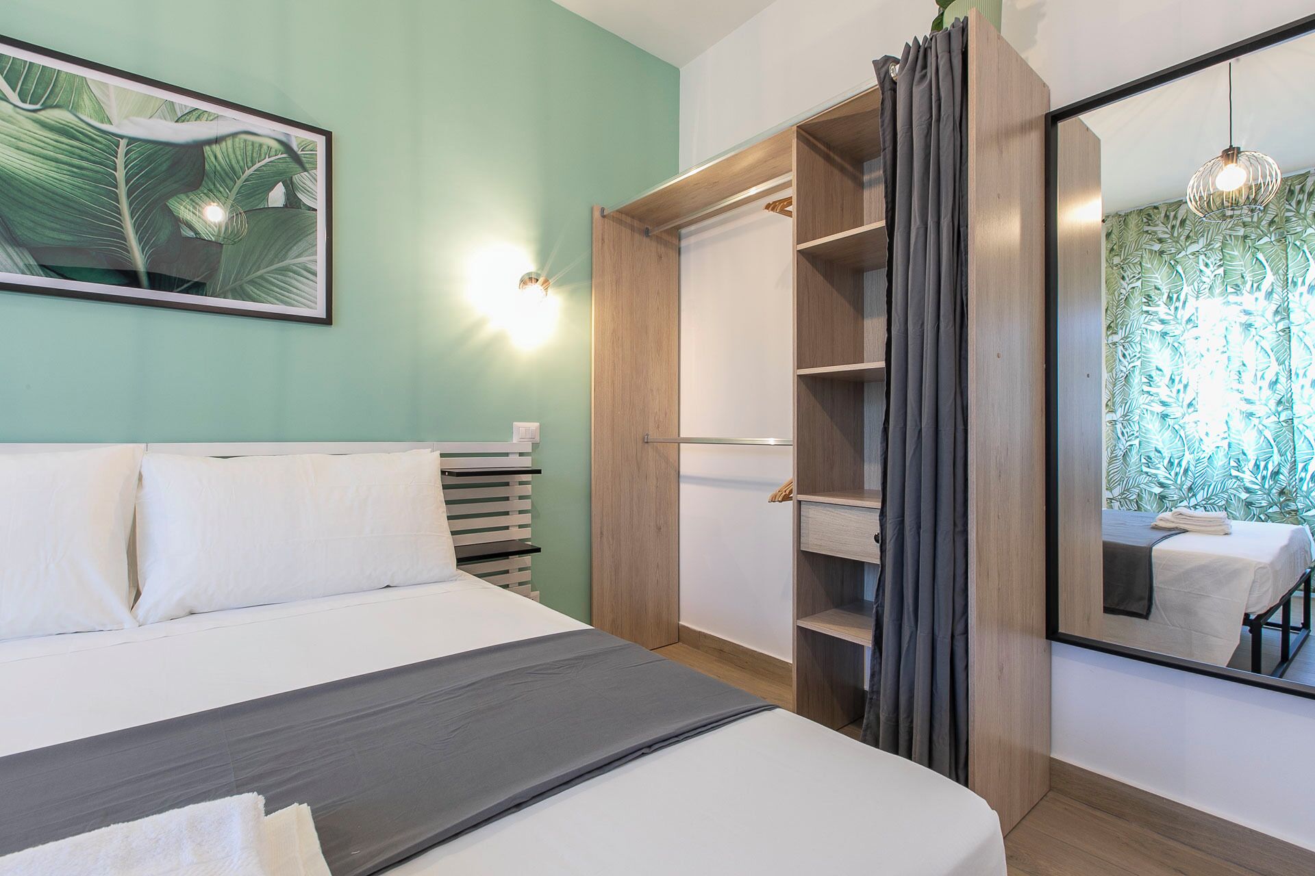 A bedroom with a bed, a wardrobe, a mirror, and a wall with a plant.