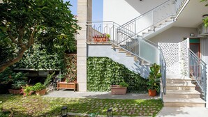 Un espacio exterior moderno con una escalera que lleva a un balcón, rodeado de abundante vegetación y plantas en macetas.