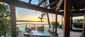 Terraza de madera con mesa y sillas de vidrio, con vista a un cuerpo de agua al atardecer.
