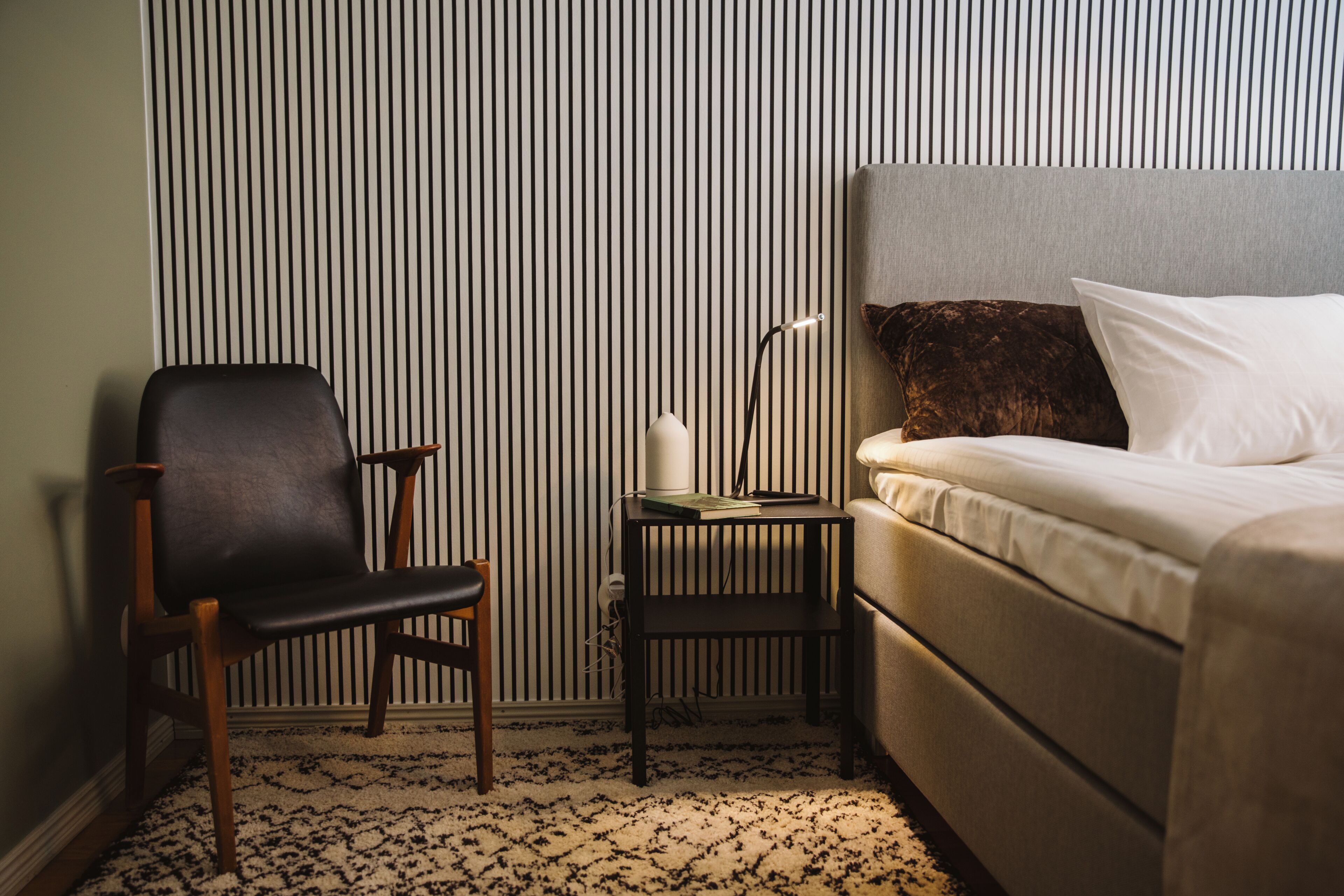 Une chambre d’hôtel avec un lit, une chaise, une petite table et un mur orné de rayures verticales.