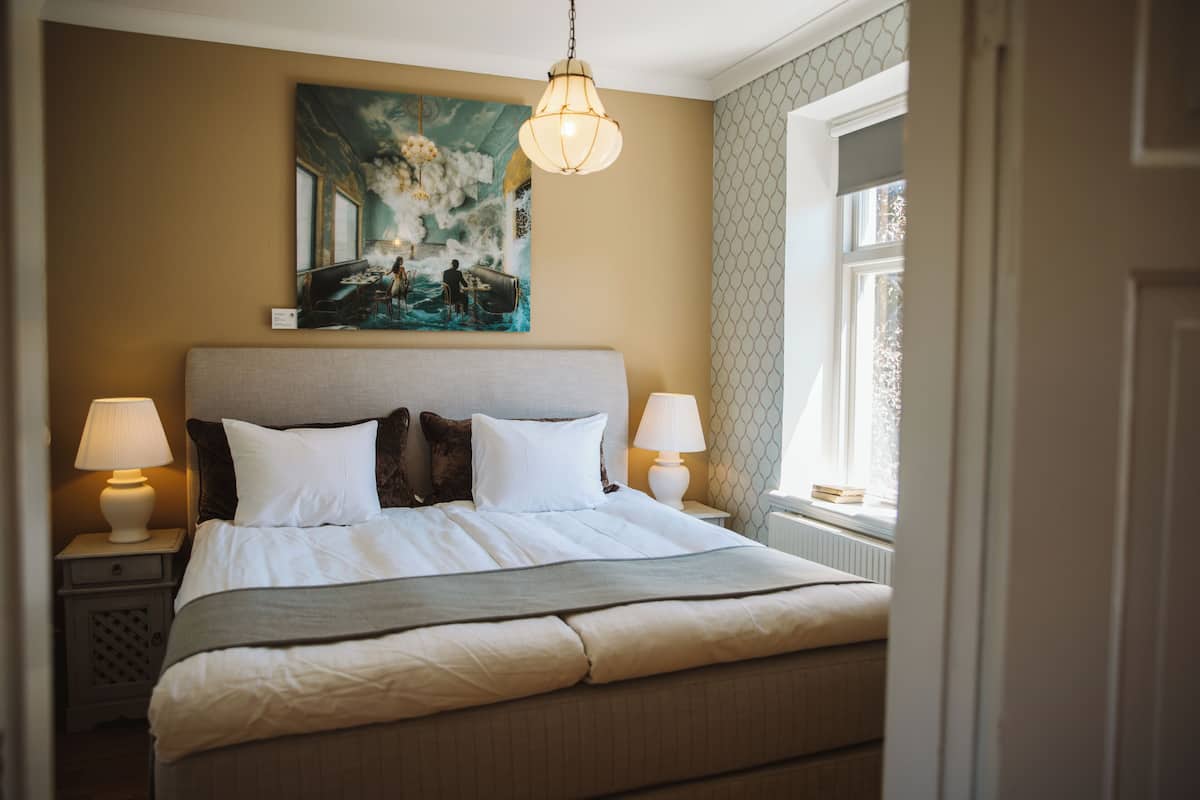 Un lit bien fait, avec du linge de lit blanc et un plaid gris, est encadré par deux lampes de chevet. Une grande peinture représentant un paysage marin est accrochée au-dessus du lit, et une fenêtre avec un rideau à motifs est visible sur la droite.