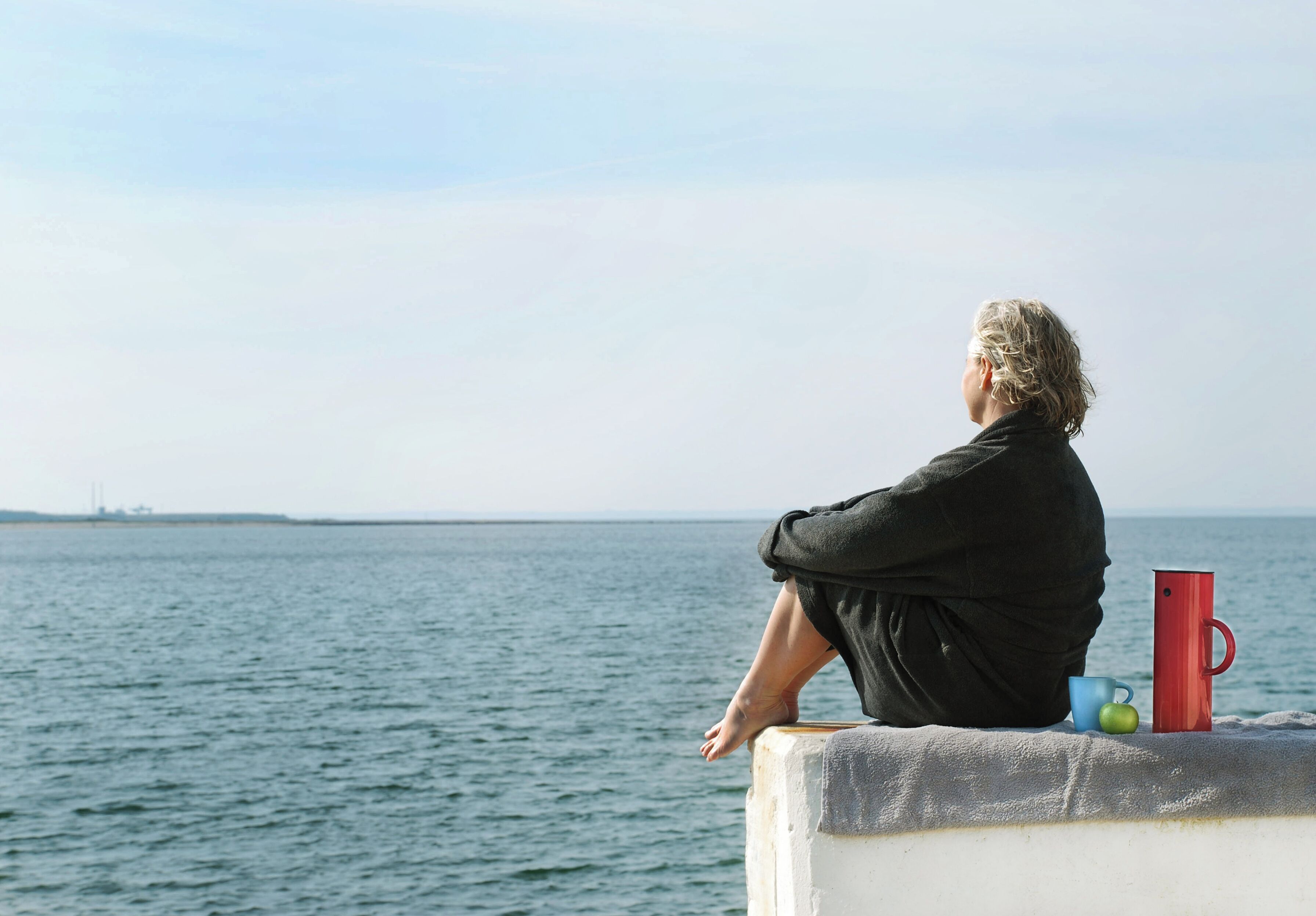 Una persona sentada sobre un borde de hormigón junto al mar, mirando al agua.