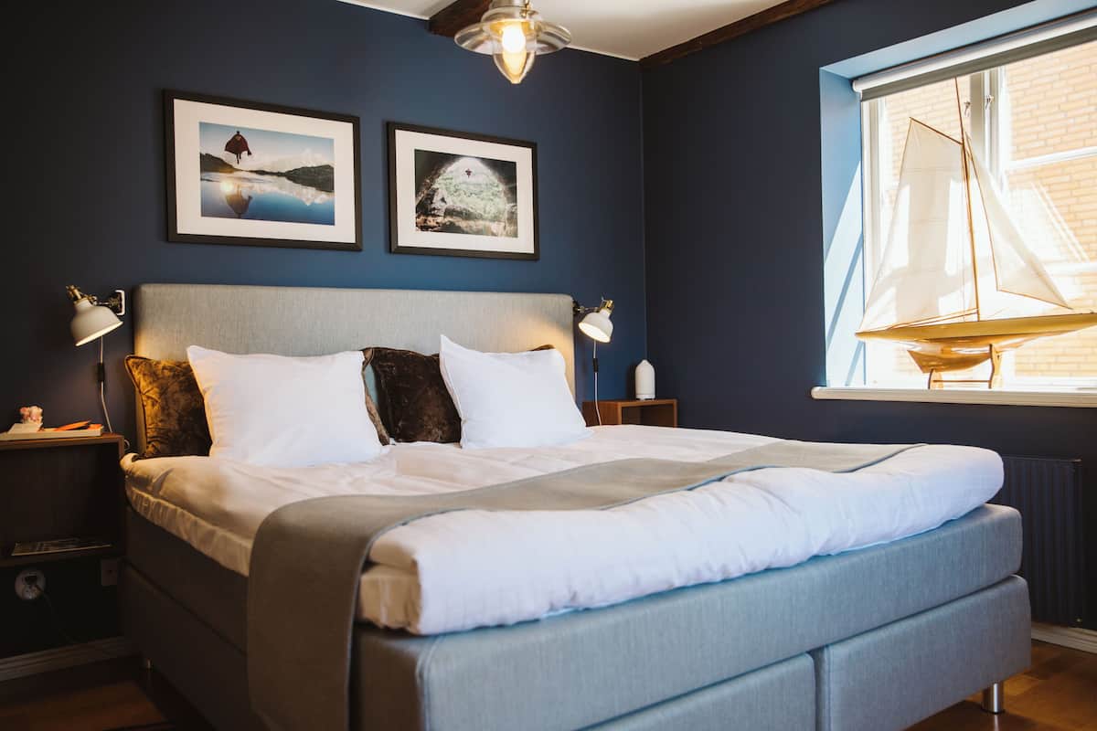 Un lit bien fait, avec du linge de lit blanc, une tête de lit grise et deux tableaux encadrés accrochés au mur.