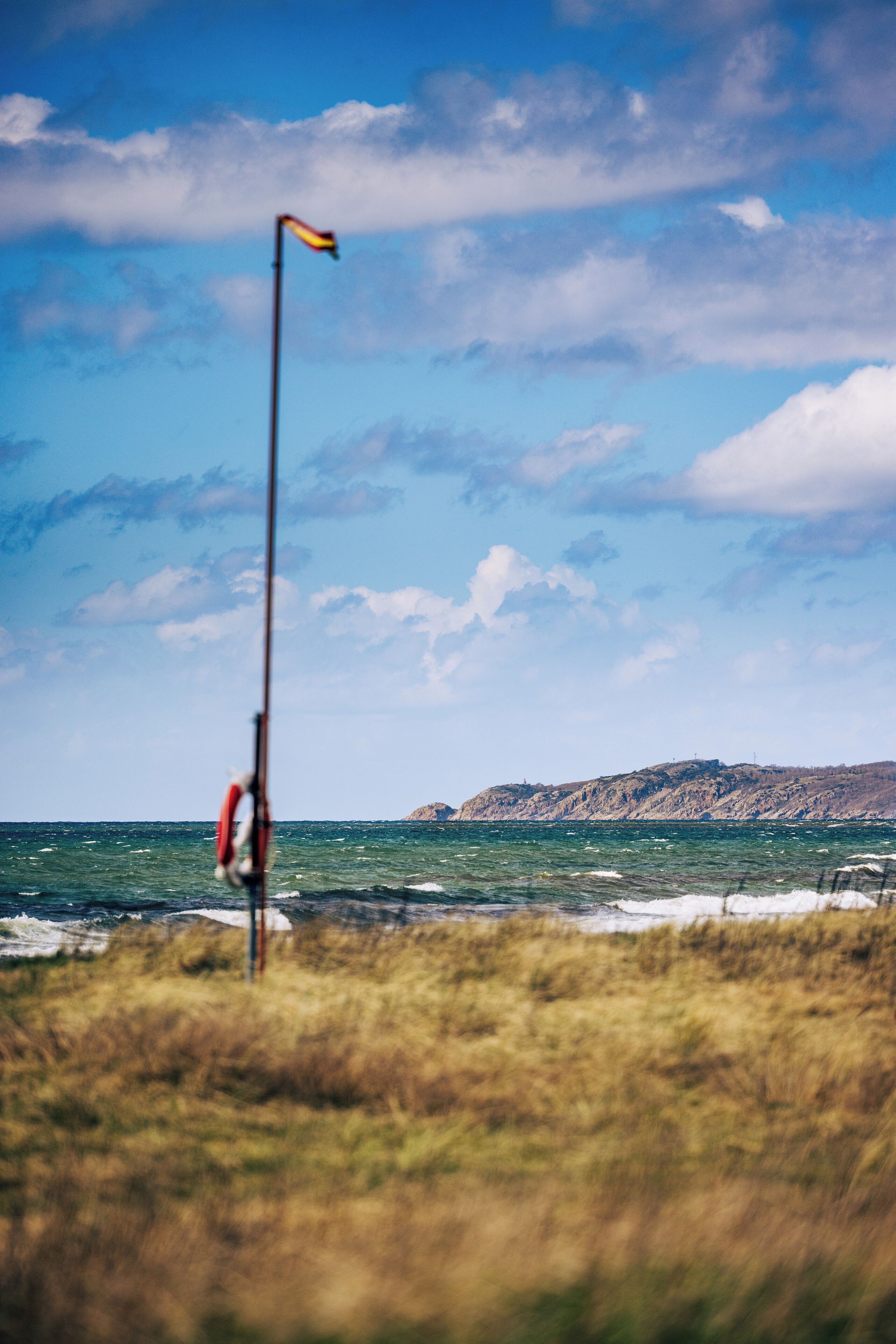 Un asta con una bandera, un primer plano con césped y una costa con una colina al fondo.