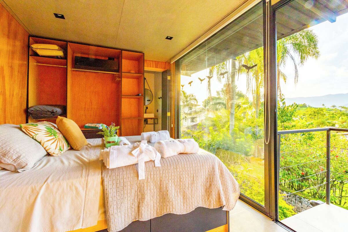 A bedroom with a large bed, wooden storage, and a balcony with a view of greenery.