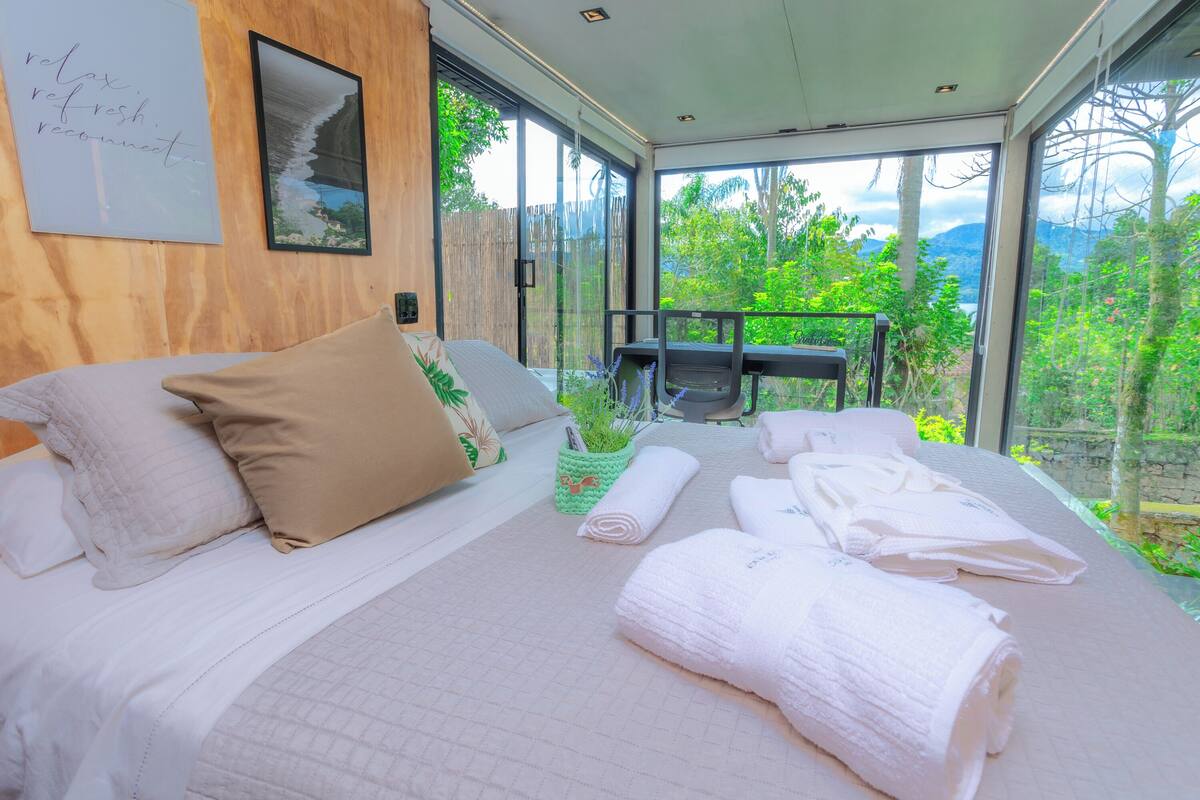A modern bedroom with a large bed, a view of greenery and mountains, and a balcony with a table and chairs.