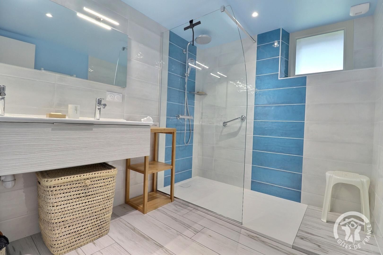 A modern bathroom with a glass-enclosed shower, a wooden chair, a small stool, and a woven basket.
