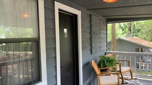 A porch with a wooden rocking chair and a small table, a door with a number sign, and a view of a tree-lined street. - Loblolly Pines Adventure Camp (Eureka Springs)