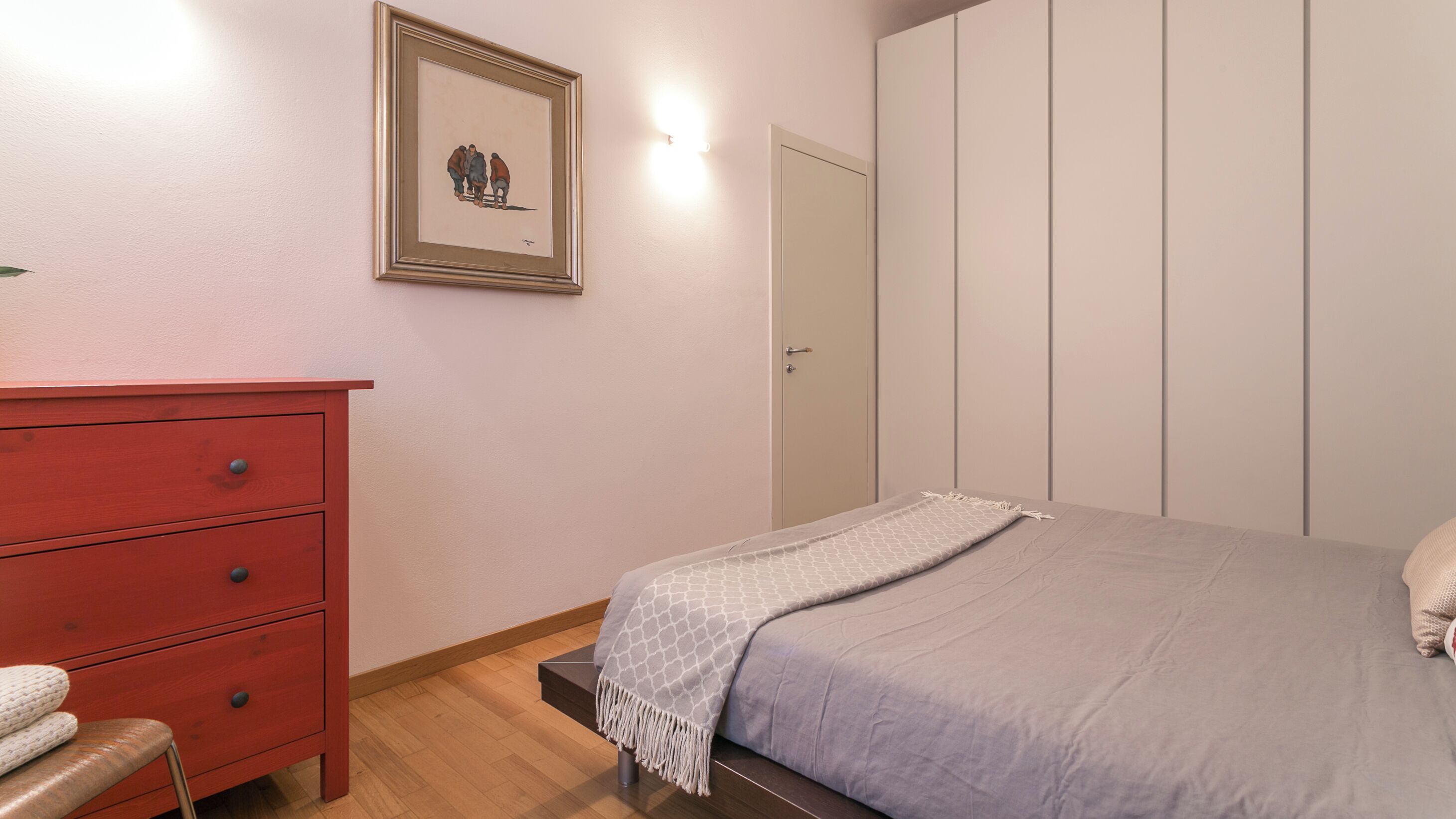A bedroom with a bed, a red dresser, a framed picture on the wall, and a door.