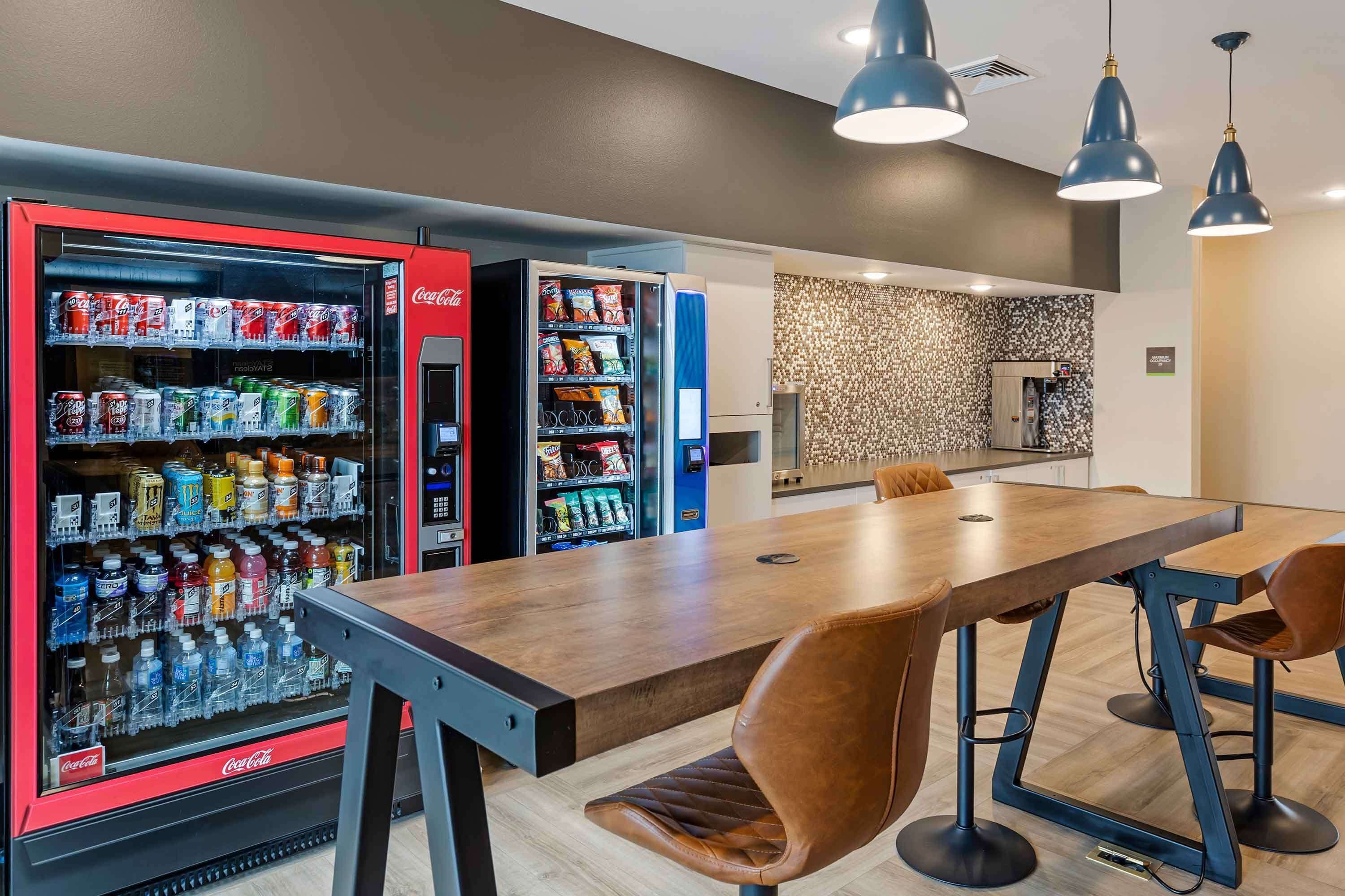 A modern dining area with a wooden table and brown leather chairs, a vending machine with snacks and drinks, and a patterned wall in the background.