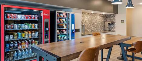 A modern dining area with a wooden table and brown leather chairs, a vending machine with snacks and drinks, and a patterned wall in the background.