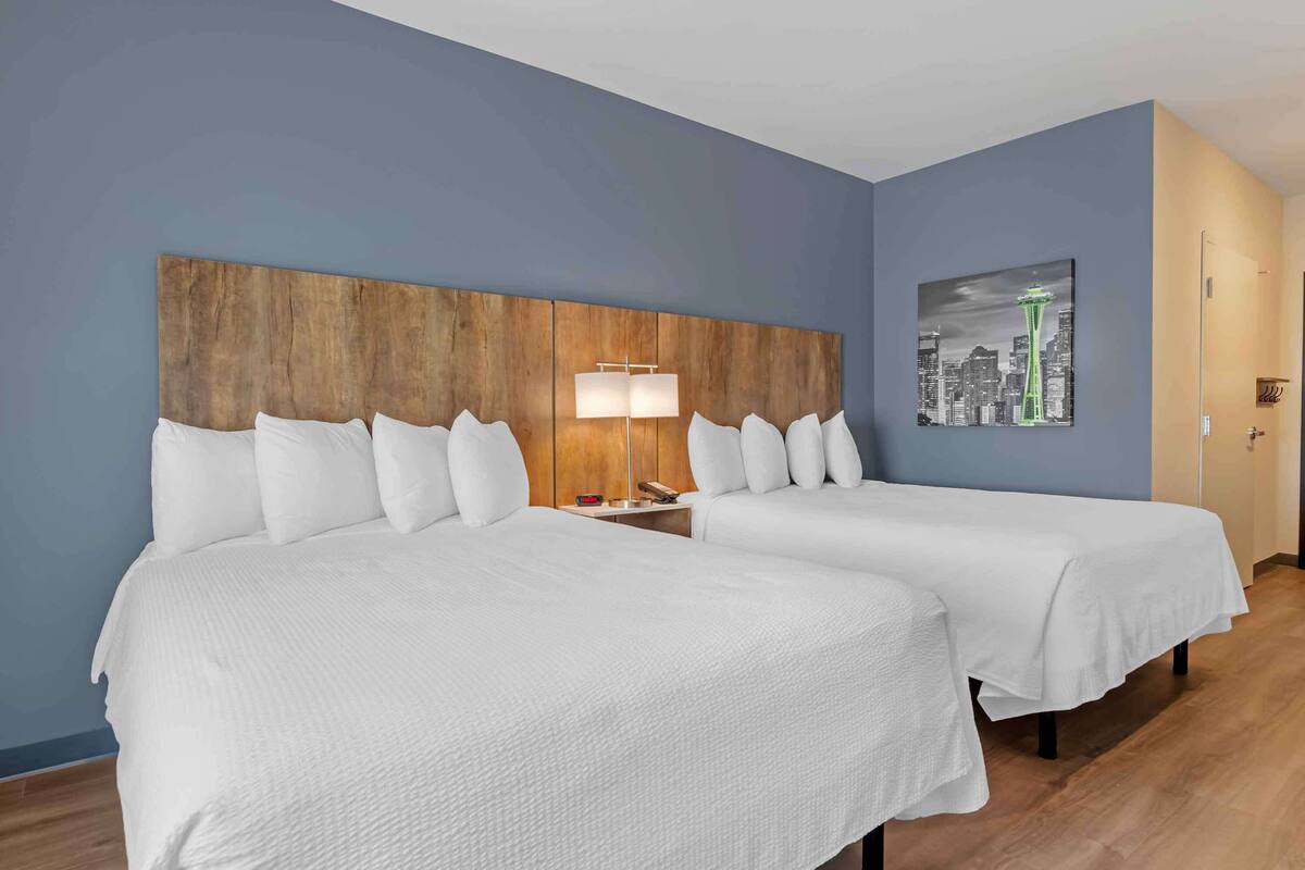 A hotel room with two beds, a wooden headboard, a cityscape painting, and a nightstand with a lamp.