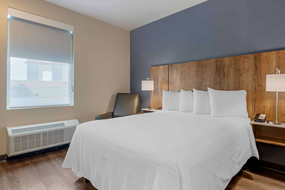 A hotel room with a large bed, a wooden headboard, two bedside lamps, a chair, and a window with blinds.