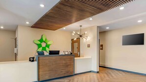 Un espace d’accueil doté d’un comptoir de réception en bois, d’une œuvre d’art abstraite verte au mur, d’un lustre moderne et d’un téléviseur à écran plat fixé au mur.