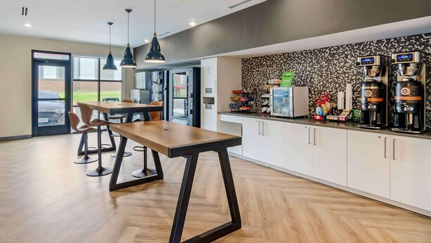 Une cuisine moderne avec une table en bois et des tabourets de bar, un coin café équipé de plusieurs distributeurs, et une vue sur l’extérieur grâce à de grandes fenêtres.
