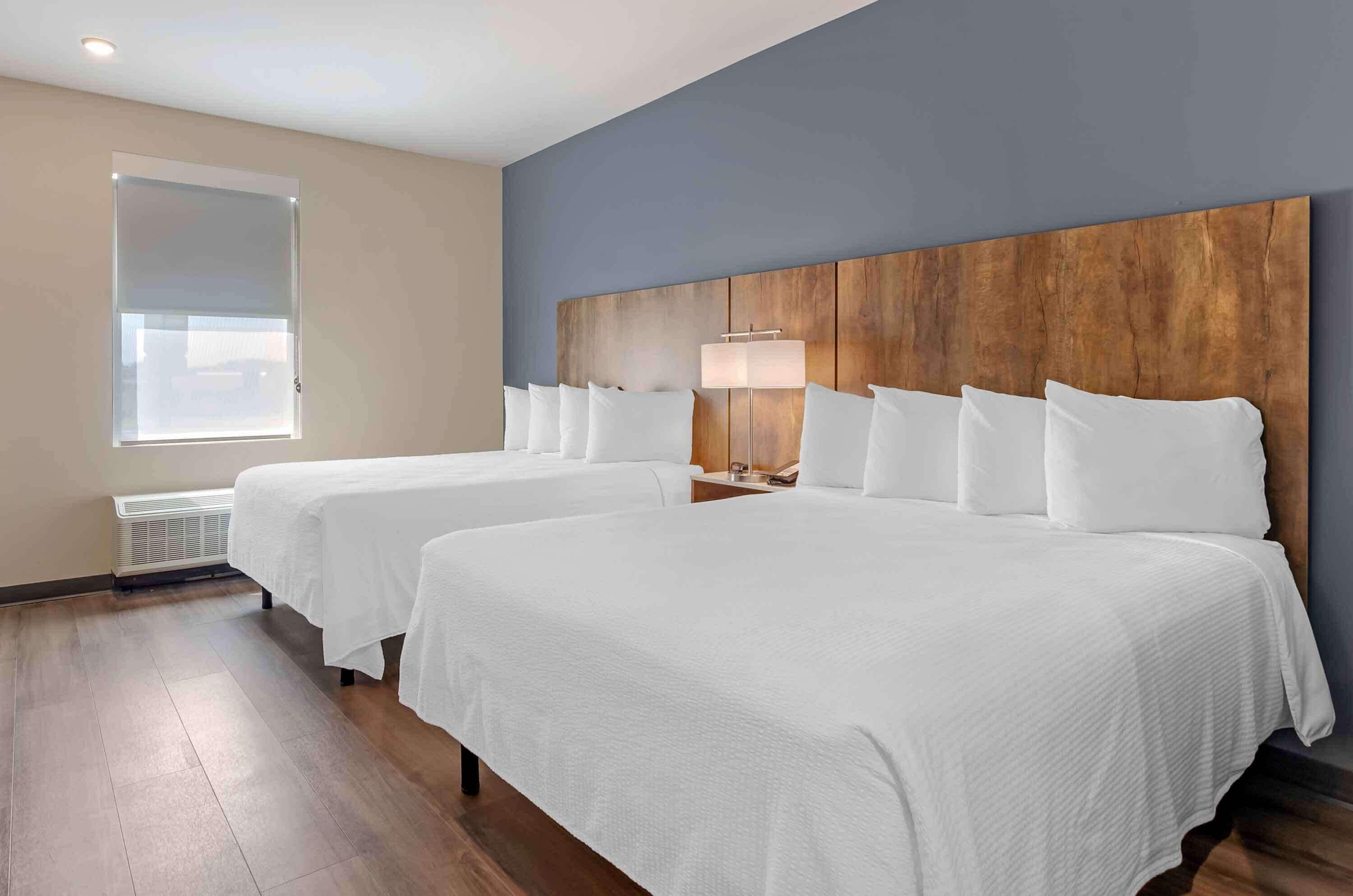 A hotel room with two beds, a wooden headboard, a bedside lamp, and a window with blinds.