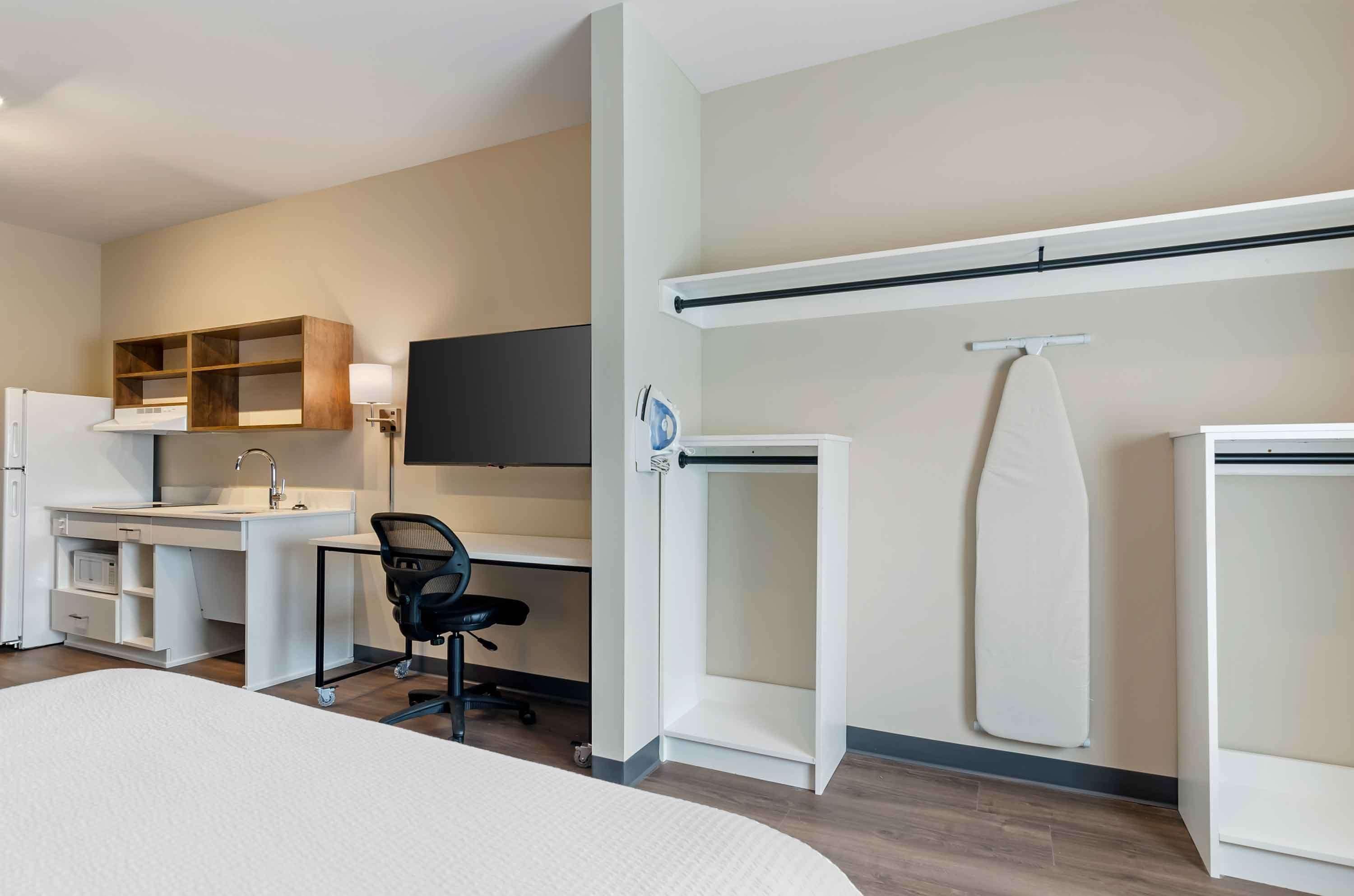 A modern hotel room with a bed, a desk with a chair, a built-in wardrobe, a flat-screen TV, and a small ironing board.